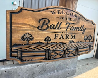 May include: A rectangular wooden sign with a dark brown stain and carved black lettering that reads Welcome to the Ball Family Farm. The design features a carved landscape with a fenced field, trees, and a small tractor silhouette.