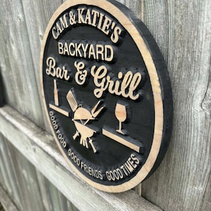 May include: A circular wooden sign with the text "CAM & KATIE'S BACKYARD Bar & Grill" carved into it. The sign includes images of grilling tools, a wine glass, and the phrases "GOOD FOOD, GOOD FRIENDS, GOOD TIMES."