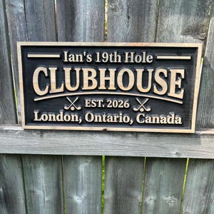 May include: A rectangular wooden sign with a black background and light brown lettering. The sign reads "Ian's 19th Hole CLUBHOUSE EST. 2026" with crossed golf clubs. Below, it says "London, Ontario, Canada".