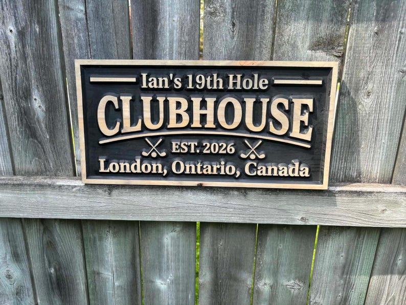May include: A rectangular wooden sign with a black background and light brown lettering. The sign reads "Ian's 19th Hole CLUBHOUSE EST. 2026" with crossed golf clubs. Below, it says "London, Ontario, Canada".