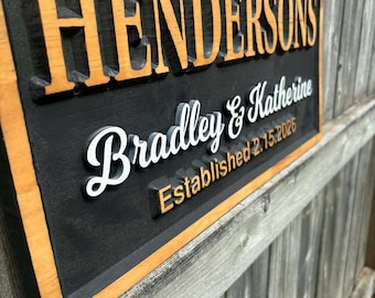 May include: A wooden sign with the name "Hendersons" in raised, light brown letters. Below, in white script, are the names "Bradley & Katherine" and the date "Established 2.15.2026". The sign has a black background and a light brown frame.