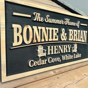 May include: A rectangular wooden sign with a black background and a light wood frame. The sign reads "The Summer Home of BONNIE & BRIAN HENRY Cedar Cove, White Lake" in a combination of fonts. Two small chair graphics are included.
