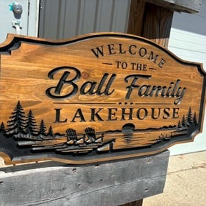 May include: A rectangular wooden sign with a carved, dark-stained finish featuring the text "WELCOME TO THE Ball Family LAKEHOUSE". The design includes a relief-carved landscape of pine trees, a lake, and two chairs along the bottom edge.