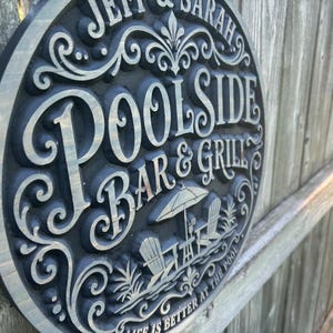 Peut inclure: Un panneau rond noir et argenté avec les mots "Jeff & Sarah" et "Poolside Bar & Grill" dans une police décorative. Le panneau présente une illustration d'une scène de plage avec des chaises, un parasol et le texte "Life is better at the pool."