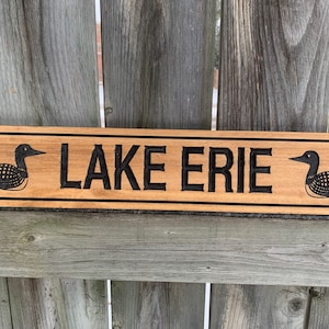 May include: A wooden sign with the words "LAKE ERIE" carved into it. The sign is decorated with two black loon birds on either side of the text.