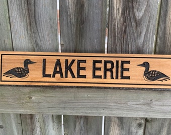 May include: A wooden sign with the words "LAKE ERIE" carved into it. The sign is decorated with two black loon birds on either side of the text.
