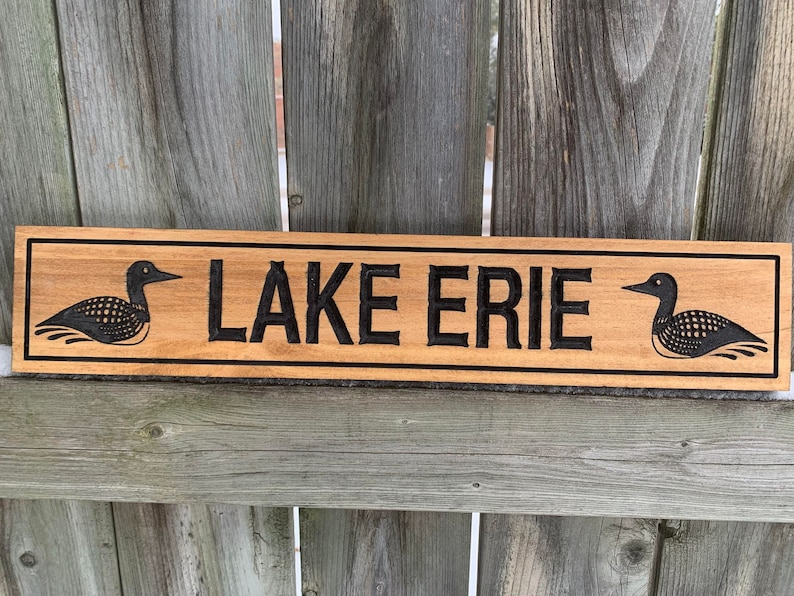 May include: A wooden sign with the words "LAKE ERIE" carved into it. The sign is decorated with two black loon birds on either side of the text.