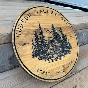 May include: A round wooden sign with the text "Hudson Valley Sauna Since 2023 Ignite Your Spirit" and a black and white illustration of a cabin in the woods.