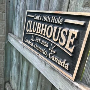 May include: A rectangular wooden sign with the text "Ian's 19th Hole CLUBHOUSE EST. 2026 London, Ontario, Canada". The sign is black with gold lettering and a gold border. Two crossed golf clubs are featured.