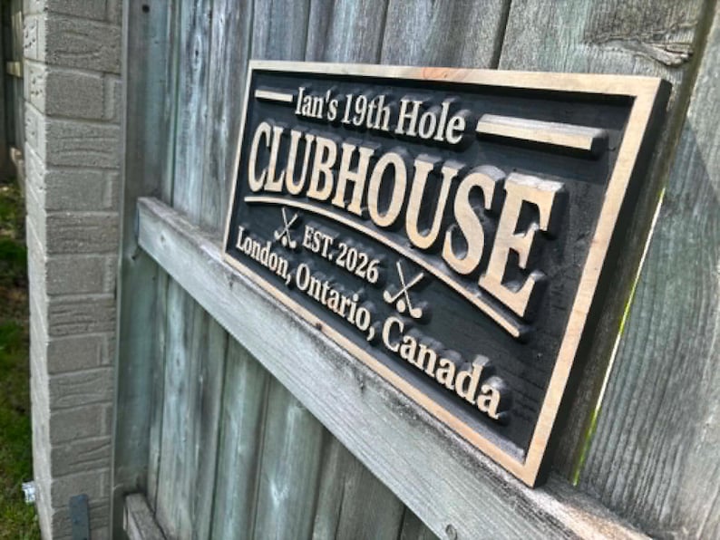 May include: A rectangular wooden sign with the text "Ian's 19th Hole CLUBHOUSE EST. 2026 London, Ontario, Canada". The sign is black with gold lettering and a gold border. Two crossed golf clubs are featured.