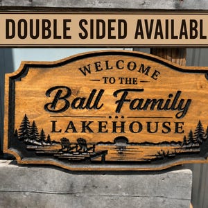 May include: A rectangular wooden sign with a dark brown finish featuring carved black lettering that reads "WELCOME TO THE Ball Family LAKEHOUSE". The design includes a carved landscape of pine trees, a dock, and chairs by a lake.