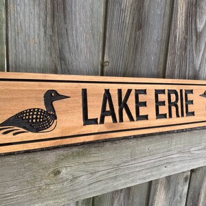 May include: A wooden sign with the words "LAKE ERIE" carved into it. The sign is decorated with two black loon birds on either side of the text.
