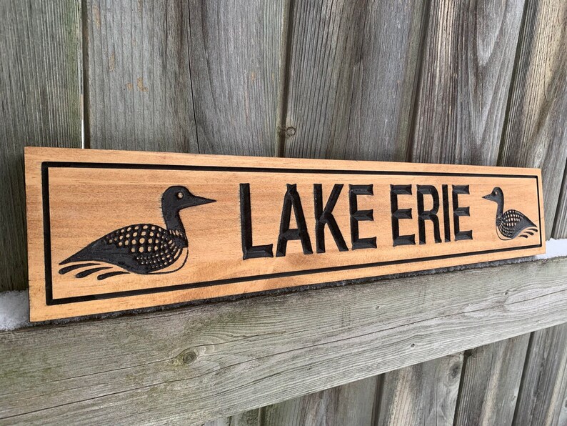 May include: A wooden sign with the words "LAKE ERIE" carved into it. The sign is decorated with two black loon birds on either side of the text.