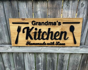 May include: Wooden sign with the words "Grandma's Kitchen" in black, along with a fork and spoon silhouette. The phrase "Homemade with Love" is also included. The sign is mounted on a wooden fence.