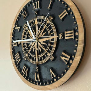 May include: A round wooden wall clock with a black face and a compass rose design in the center. The clock features Roman numerals and hands indicating the time. The outer rim is a natural wood color, and the clock has a nautical theme.