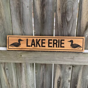 May include: Wooden sign with the text "LAKE ERIE" in black lettering. The sign is decorated with two black loon birds on either side of the text.