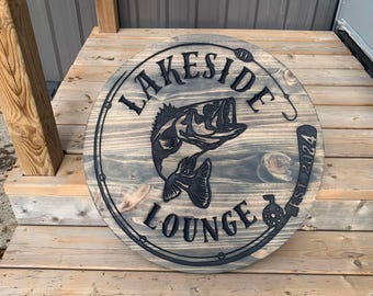 May include: A round wooden sign with the text "Lakeside Lounge Est 2024" and a black silhouette of a bass fish. The sign has a distressed wood finish and a rope border.