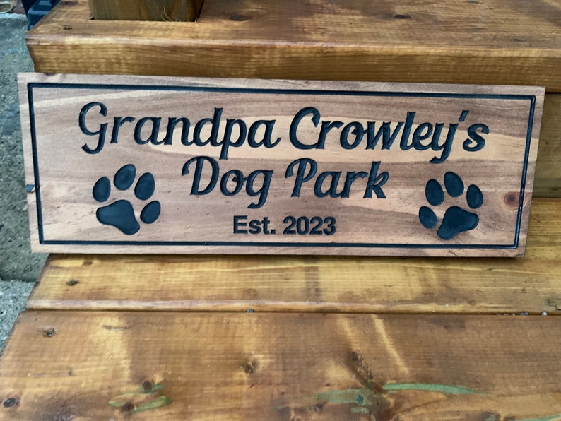 May include: A wooden sign with the text "Grandpa Crowley's Dog Park Est. 2023" and two black paw prints.