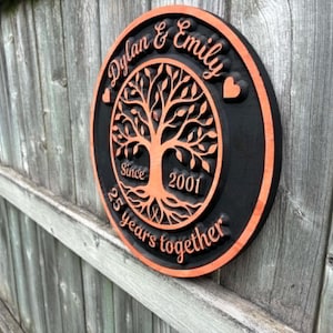 May include: A circular wooden plaque with a black base and orange edging. It displays the names "Dylan & Emily" and a tree motif. The text reads "Since 2001" and "25 years together."