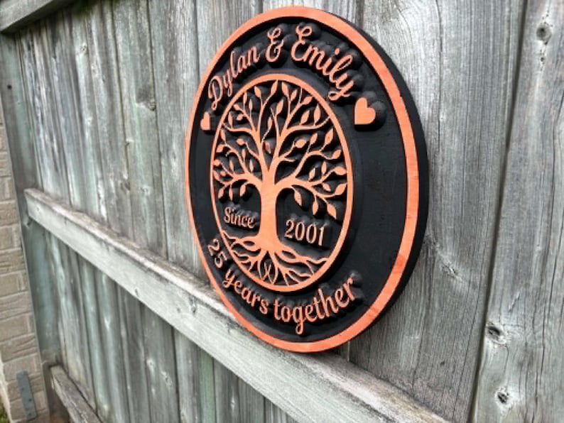 May include: A round, wooden plaque with a black background and orange trim. The plaque features the names "Dylan & Emily" and a tree design. Text includes "Since 2001" and "25 years together."