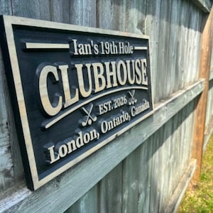 May include: A rectangular wooden sign with a black background and light wood trim. The sign reads "Ian's 19th Hole CLUBHOUSE EST. 2026" with crossed golf clubs. The text "London, Ontario, Canada" is also displayed.
