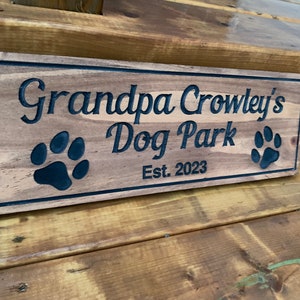 May include: A wooden sign with the text "Grandpa Crowley's Dog Park Est. 2023" in black lettering. Two black paw prints are on either side of the text.