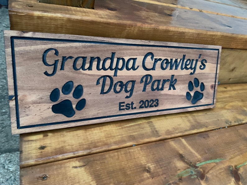 May include: A wooden sign with the text "Grandpa Crowley's Dog Park Est. 2023" in black lettering. Two black paw prints are on either side of the text.