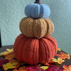 May include: A stack of three crocheted pumpkins in shades of blue, orange, and rust. The pumpkins are arranged vertically, with the largest at the bottom and the smallest at the top. Brown stem tops the top pumpkin. Colorful fall leaves surround the base.