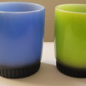 Puede incluir: Dos tazas de vidrio vintage. Una es azul, la otra verde. Ambas tienen una base negra y acanalada y un asa. Ideales para café, té u otras bebidas.