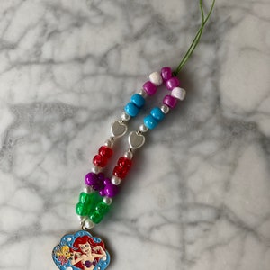 May include: A colorful beaded lanyard featuring a pendant with a cartoon image of Ariel from The Little Mermaid. The lanyard has a variety of beads in colors like red, purple, green, blue, white, and pink. Silver heart-shaped beads are also included.