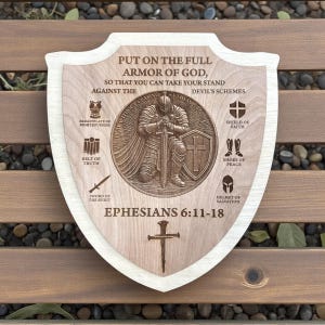 May include: Wooden shield-shaped plaque with a light wood border. The plaque features engraved text and illustrations of armor, including a helmet, shield, and sword. The text reads "PUT ON THE FULL ARMOR OF GOD" and "EPHESIANS 6:11-18".