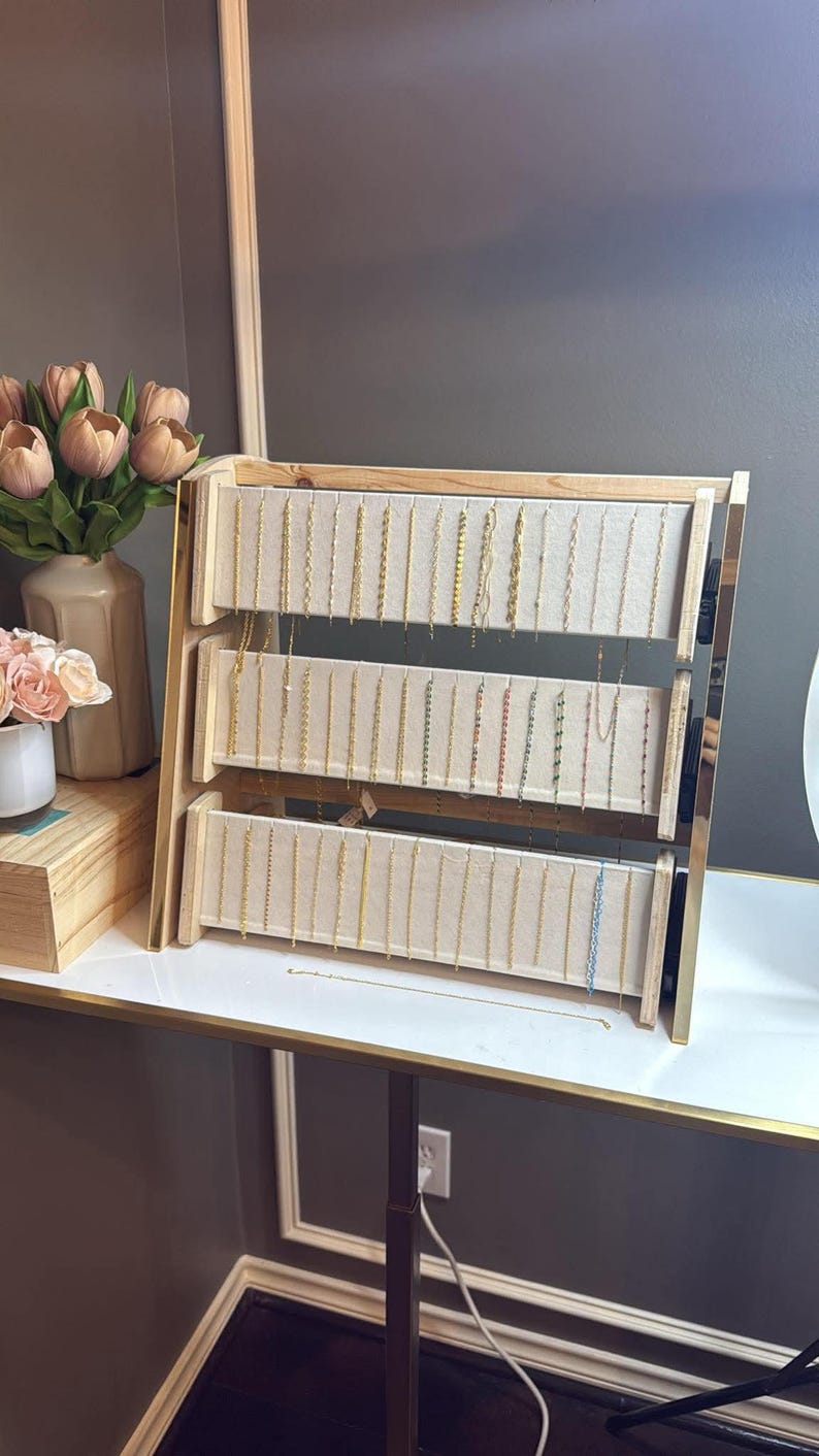 May include: A three-tiered jewelry display stand with a light wood frame and beige fabric backing. Various necklaces in gold and silver tones hang from the display. The stand is on a white surface with a gold trim, and a vase of flowers is visible in the background.