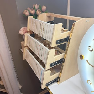 May include: A wooden jewelry organizer with three pull-out drawers. Each drawer has multiple rows of hanging space for necklaces and bracelets. The organizer has a light wood finish with gold trim and is set against a neutral background.