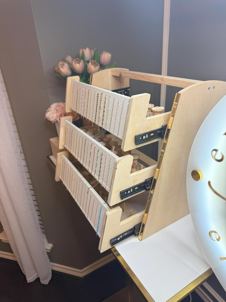 May include: A wooden jewelry organizer with three pull-out drawers. Each drawer has multiple rows of hanging space for necklaces and bracelets. The organizer has a light wood finish with gold trim and is set against a neutral background.