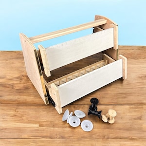 May include: A wooden craft organizer with two pull-out drawers, a handle, and various small parts. The organizer is made of light-colored wood and has a natural finish. The drawers have a white fabric lining. The parts include white discs, black clips, and wooden spools.