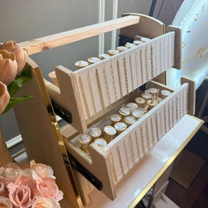 May include: A wooden jewelry display stand with two pull-out drawers. The drawers are lined with beige fabric and hold various gold necklaces and bracelets. The stand is on a white surface with gold trim.