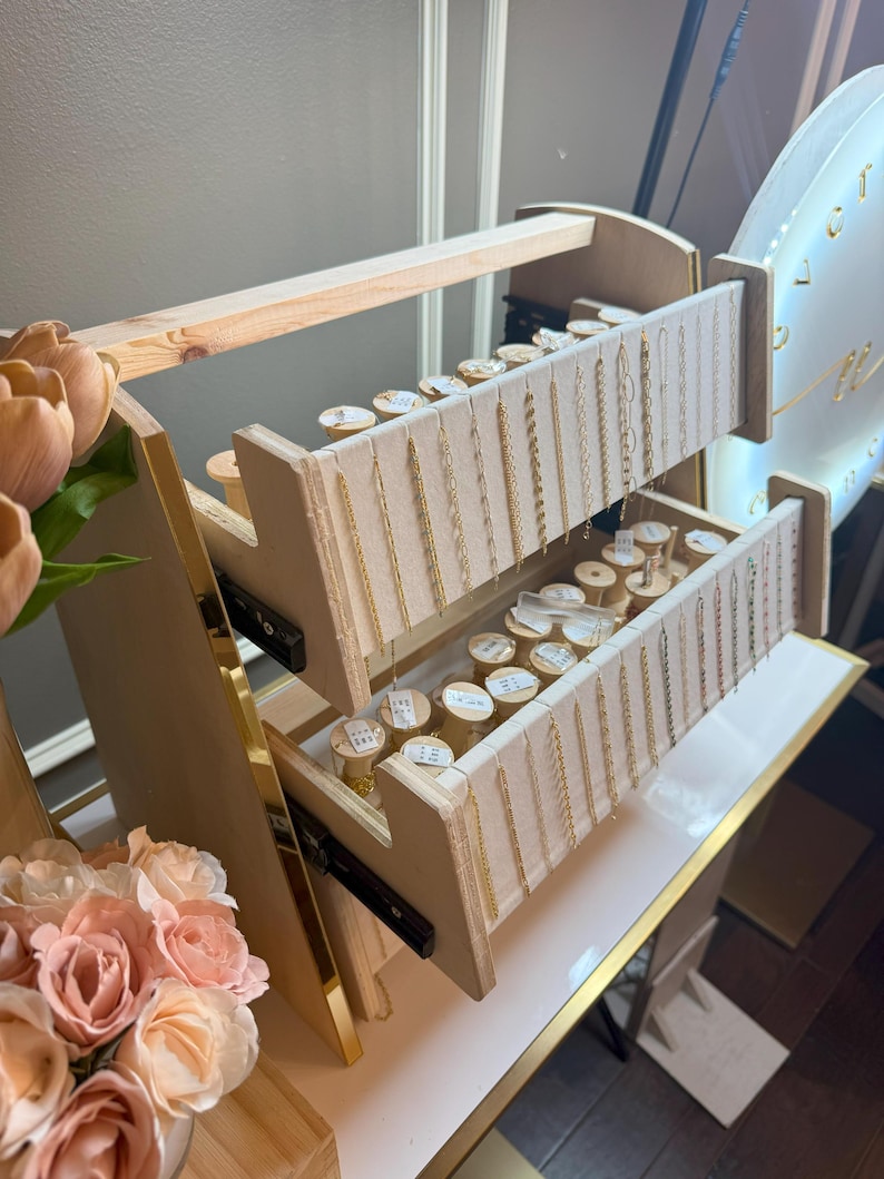 May include: A wooden jewelry display stand with two pull-out drawers. The drawers are lined with beige fabric and hold various gold necklaces and bracelets. The stand is on a white surface with gold trim.