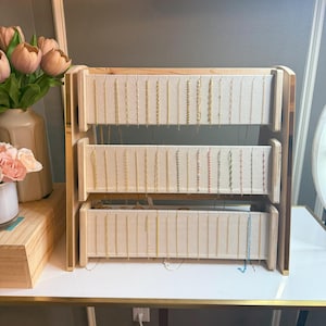 May include: A three-tiered jewelry display stand made of light wood and beige fabric. The stand holds numerous necklaces of varying lengths and colors. The stand is on a white table with a gold trim.