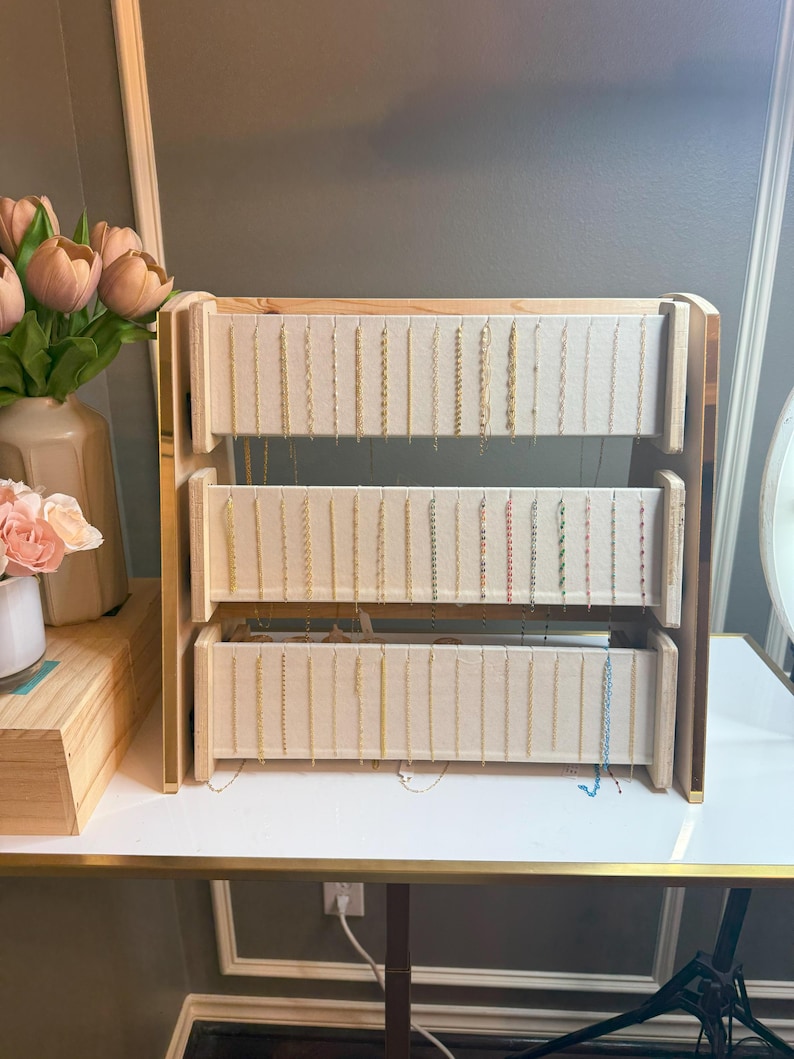 May include: A three-tiered jewelry display stand made of light wood and beige fabric. The stand holds numerous necklaces of varying lengths and colors. The stand is on a white table with a gold trim.