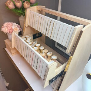 May include: A wooden jewelry display stand with multiple levels. The top level displays gold necklaces hanging from a white fabric. The bottom level contains spools of thread and more necklaces. Pink flowers in a vase are in the background.