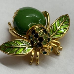 May include: A gold-toned insect brooch featuring a large, smooth, dark green cabochon body, a thorax encrusted with small black and light green crystals, and two wings detailed with bright green enamel and gold veins, plus small green crystal eyes.
