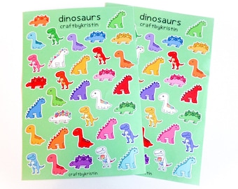 Op de afbeelding: Twee vellen met kleurrijke dinosaurus stickers op een groene achtergrond. De stickers tonen verschillende dinosaurussen in verschillende kleuren en poses. De tekst "dinosaurs craftbykristin" staat op elk vel gedrukt.