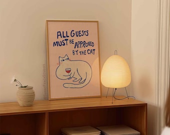 May include: A framed print with a pink background and a white cat illustration. The cat is holding a glass of wine and the text reads "ALL GUESTS MUST BE APPROVED BY THE CAT". The print is hanging on a wall above a wooden shelf with a lamp and books.