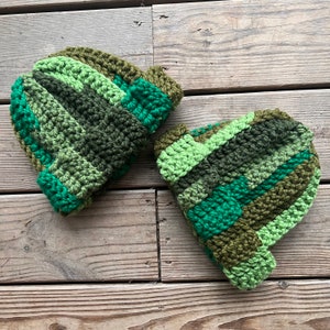 May include: Two handmade crochet beanies in shades of green, olive, and brown. The beanies feature a striped pattern with varying widths of color blocks. The hats are displayed on a weathered wooden surface, showcasing the texture of the yarn.