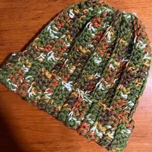 May include: A crocheted beanie hat in green, orange, white, and brown yarn. The hat has a ribbed texture and a slouchy fit.