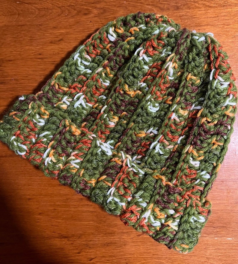 May include: A crocheted beanie hat in green, orange, white, and brown yarn. The hat has a ribbed texture and a slouchy fit.