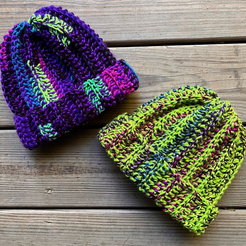 May include: Two crocheted hats, one in shades of purple and green, the other in shades of green and black.