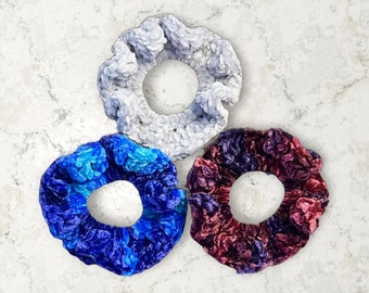 May include: Three fabric scrunchies in different colours and patterns. One is white with a fluffy texture, one is blue with a swirl pattern, and one is purple with a swirl pattern.
