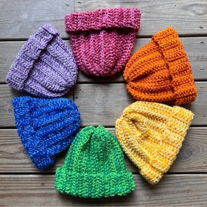 May include: Six crocheted hats in a rainbow of colors, including purple, blue, green, yellow, orange, and pink. The hats are arranged in a circle on a wooden surface.