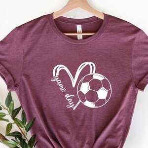 May include: Burgundy t-shirt with a white soccer ball and the text "game day" in a heart shape.